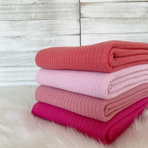 May include: Four waffle weave fabric swatches in shades of pink, coral, and fuchsia. The fabric is folded and stacked on a white faux fur surface.
