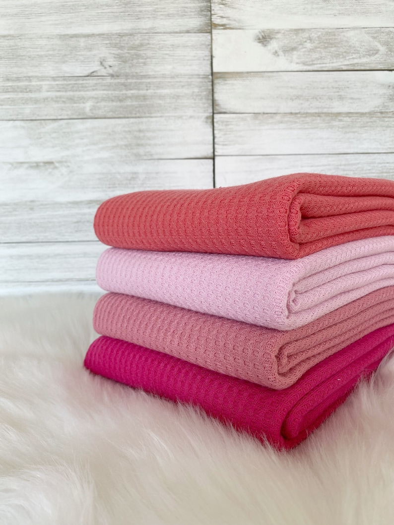 May include: Four waffle weave fabric swatches in shades of pink, coral, and fuchsia. The fabric is folded and stacked on a white faux fur surface.