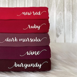 May include: Five fabric swatches stacked on top of each other. The swatches are different shades of red, from a bright red to a deep burgundy. The swatches are labeled with the following names: new red, ruby, dark marsala, wine, burgundy.