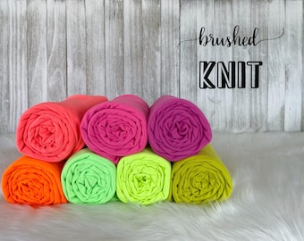 May include: Six rolls of brushed knit fabric in bright colors: neon pink, orange, green, lime green, yellow, and magenta. The fabric is rolled up and stacked on a white fluffy surface. The text "brushed knit" is written in black above the fabric.