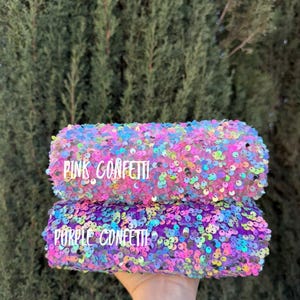 May include: Two sequined rolls, one pink and one purple, are held up against a backdrop of green foliage. The pink roll has the text "PINK CONFETTI" and the purple roll has the text "PURPLE CONFETTI". The sequins are iridescent.