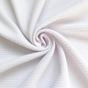May include: Close-up of white fabric with a textured, quilted pattern. The fabric is draped in soft folds, creating a swirling effect. The material appears to be lightweight and suitable for various sewing projects.