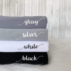 May include: Four fabric swatches stacked on top of each other. The swatches are gray, silver, white, and black. Each swatch has the color name written in white cursive on the fabric.