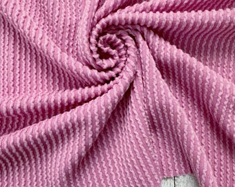 May include: Pink textured fabric with a ribbed pattern. The fabric is folded and draped on a wooden surface.