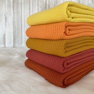 May include: A stack of five waffle weave fabric swatches in shades of yellow, orange, brown, and red. The fabric is folded and stacked on a white fluffy surface.