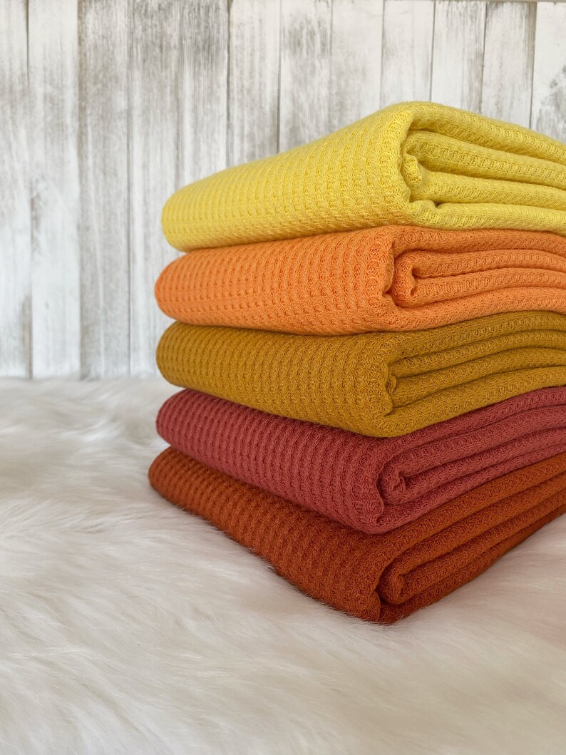 May include: A stack of five waffle weave fabric swatches in shades of yellow, orange, brown, and red. The fabric is folded and stacked on a white fluffy surface.