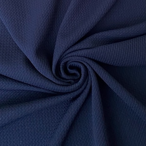 May include: Close-up of a navy blue fabric with a textured, dimpled surface. The fabric is arranged in a spiral, showcasing its drape and the play of light and shadow across its surface. The material appears soft and pliable.