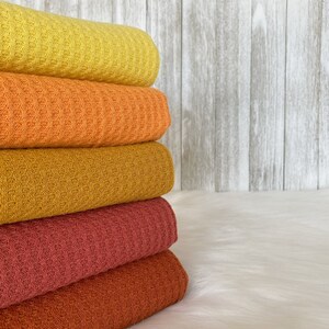 May include: A stack of five waffle weave blankets in shades of yellow, orange, brown, and red. The blankets are folded and stacked on a white surface.