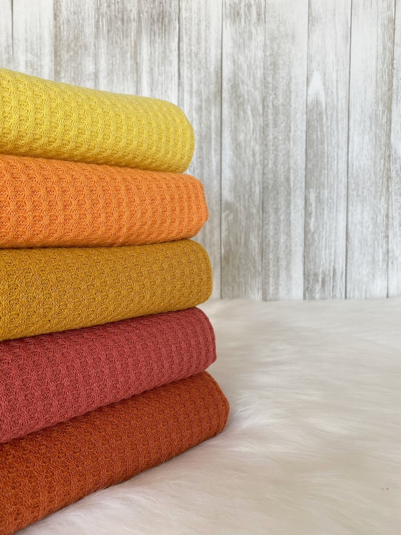May include: A stack of five waffle weave blankets in shades of yellow, orange, brown, and red. The blankets are folded and stacked on a white surface.
