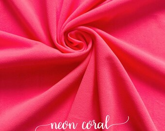 Puede incluir: Una tela de color coral neón brillante, doblada y exhibida sobre una superficie de madera. La tela es suave y lisa, con un ligero brillo. El texto "coral neón" está escrito en una fuente cursiva sobre la tela.