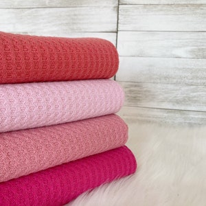 May include: Four waffle weave fabric swatches in shades of pink, coral, and fuchsia. The fabric is folded and stacked on a white fluffy surface.