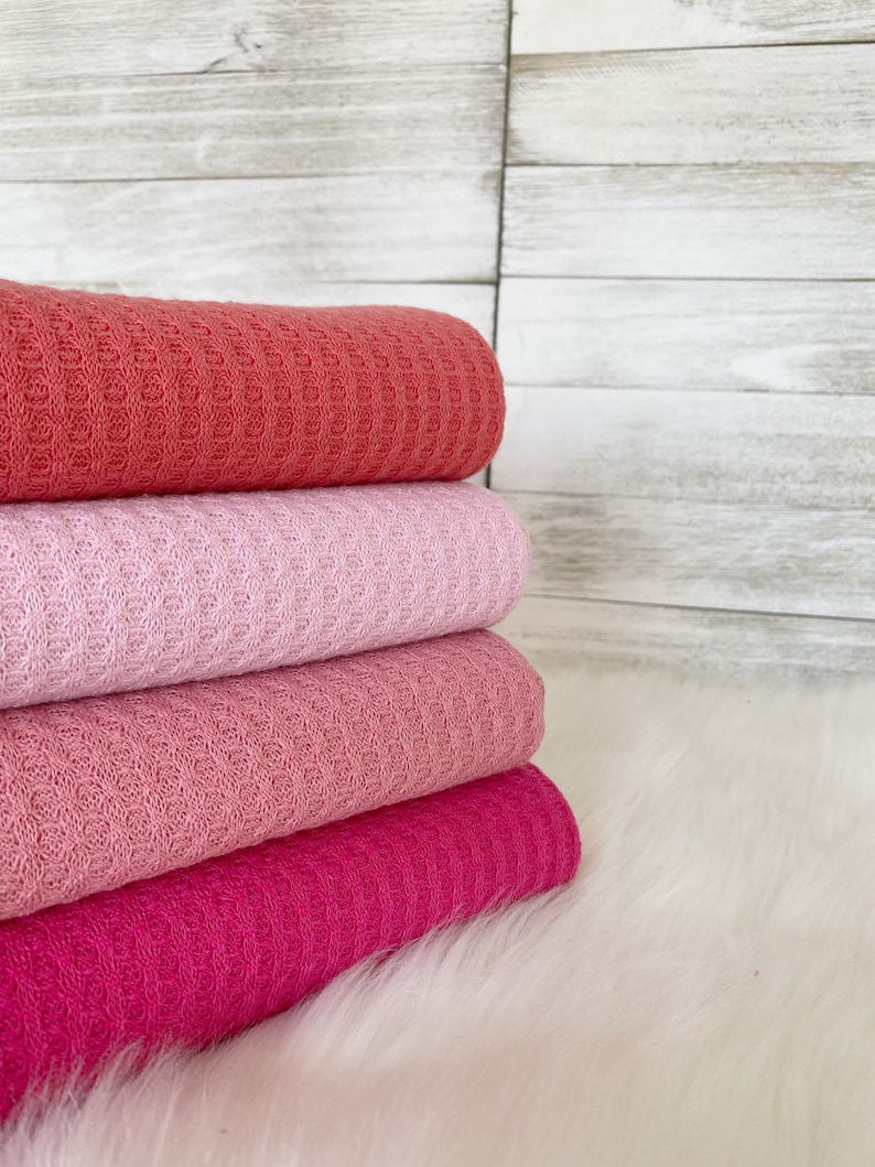 May include: Four waffle weave fabric swatches in shades of pink, coral, and fuchsia. The fabric is folded and stacked on a white fluffy surface.