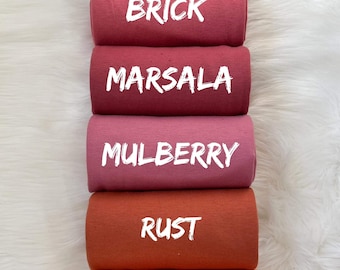 Può includere: Una pila di sei rotoli di tessuto in vari colori, tra cui bordeaux, mattone, marsala, gelso, ruggine e arancione polveroso. Ogni rotolo ha un'etichetta di testo bianca che indica il nome del colore. I rotoli sono disposti verticalmente su uno sfondo bianco e soffice.