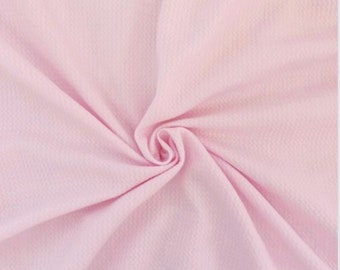 May include: Close-up of a light pink fabric with a textured surface. The fabric is gathered in a spiral pattern, creating soft folds and shadows. The overall appearance is smooth and delicate, suggesting a textile.