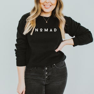 Puede incluir: Una sudadera negra con la palabra "NOMAD" impresa en blanco en la parte delantera. La modelo lleva pantalones vaqueros negros y un collar de oro.