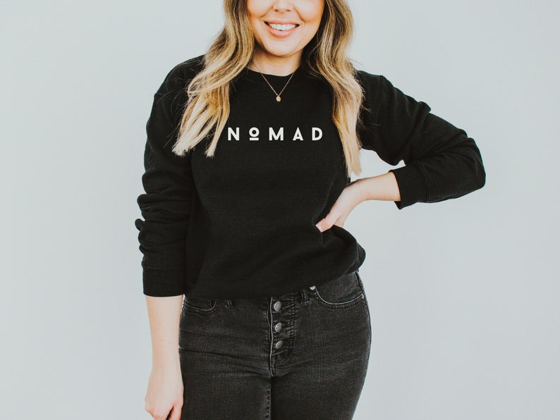 Puede incluir: Una sudadera negra con la palabra "NOMAD" impresa en blanco en la parte delantera. La modelo lleva pantalones vaqueros negros y un collar de oro.