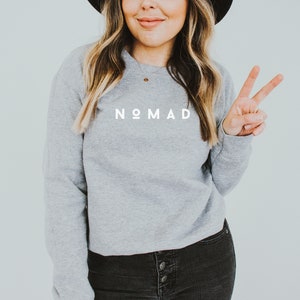 Puede incluir: Una sudadera gris con la palabra "NOMAD" impresa en blanco en la parte delantera. La modelo lleva pantalones vaqueros negros y un sombrero negro.