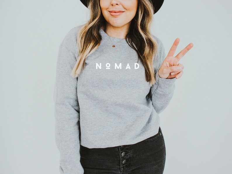 Puede incluir: Una sudadera gris con la palabra "NOMAD" impresa en blanco en la parte delantera. La modelo lleva pantalones vaqueros negros y un sombrero negro.