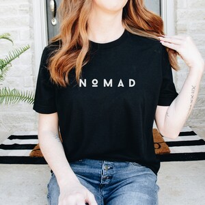 May include: A black t-shirt with the word "NOMAD" printed in white on the front. The shirt is being worn by a person with long brown hair.