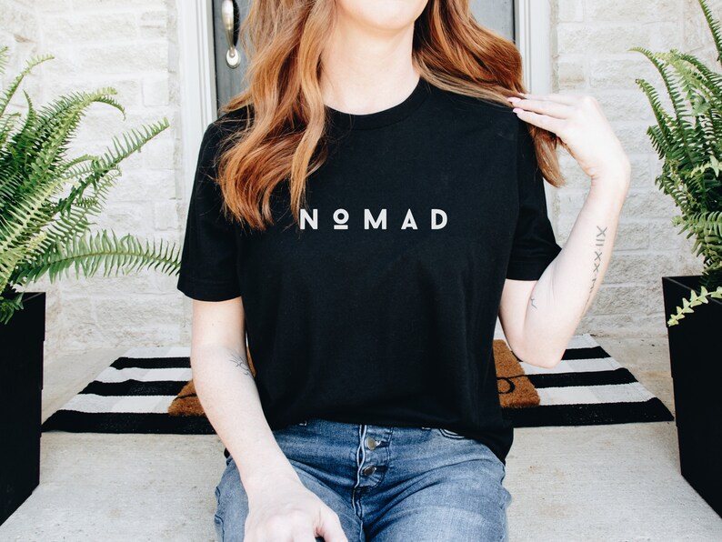 May include: A black t-shirt with the word "NOMAD" printed in white on the front. The shirt is being worn by a person with long brown hair.