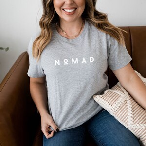 May include: A woman wearing a gray t-shirt with the word "NOMAD" printed in white on the front. She is sitting on a brown leather couch with a beige pillow.