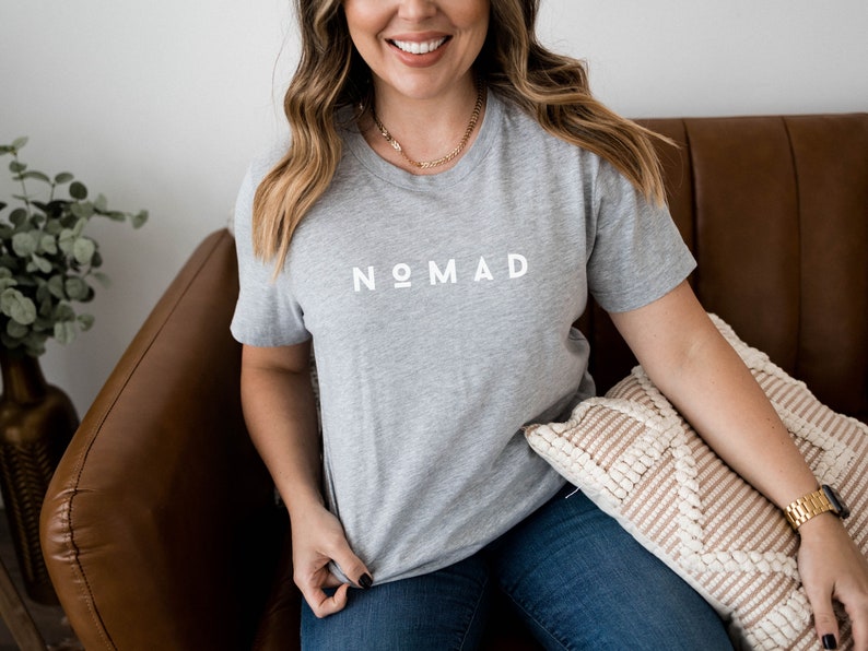May include: A woman wearing a gray t-shirt with the word "NOMAD" printed in white on the front. She is sitting on a brown leather couch with a beige pillow.