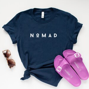 May include: A navy blue t-shirt with the word "NOMAD" printed in white on the front. The shirt is laid flat on a white surface with a pair of brown sunglasses and a pair of purple sandals.