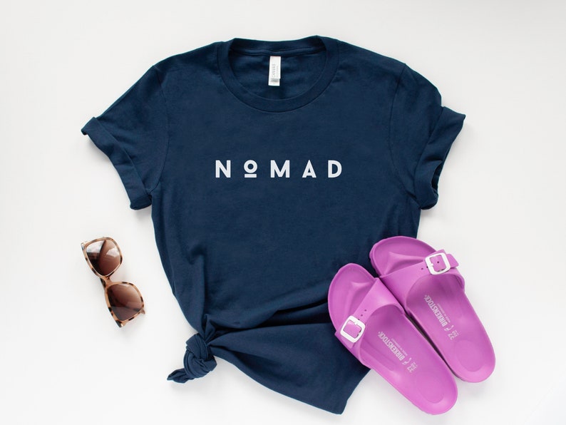 May include: A navy blue t-shirt with the word "NOMAD" printed in white on the front. The shirt is laid flat on a white surface with a pair of brown sunglasses and a pair of purple sandals.