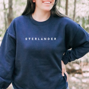 May include: A navy blue sweatshirt featuring the word "OVERLANDER" in white lettering. The sweatshirt has a crew neck and long sleeves. The image shows a person wearing the sweatshirt.