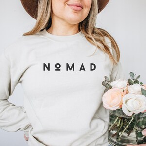 Puede incluir: Una sudadera de color beige claro con la palabra "NOMAD" impresa en negro en la parte delantera. La sudadera la est&aacute; sujetando una persona que tambi&eacute;n sostiene un jarr&oacute;n de rosas blancas y rosas.
