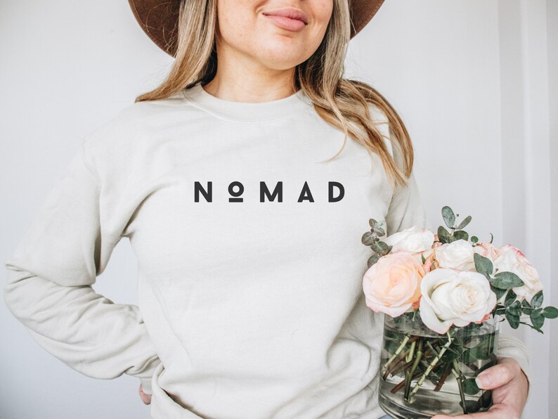 Puede incluir: Una sudadera de color beige claro con la palabra "NOMAD" impresa en negro en la parte delantera. La sudadera la est&aacute; sujetando una persona que tambi&eacute;n sostiene un jarr&oacute;n de rosas blancas y rosas.