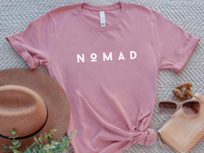 May include: A dusty rose colored t-shirt with the word "NOMAD" printed in white on the front.