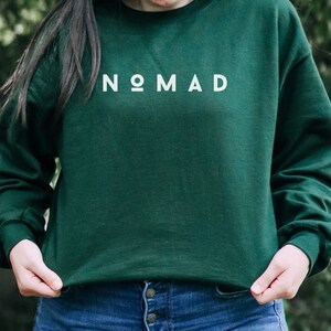 Puede incluir: Una sudadera verde oscuro con la palabra "NOMAD" impresa en blanco en la parte delantera.