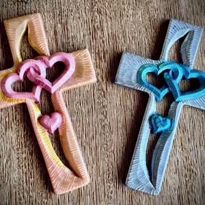 May include: Two decorative crosses featuring textured surfaces and carved details. One cross is light orange with pink interlocking hearts and a small pink heart below. The other cross is light blue with teal interlocking hearts and a small teal heart.