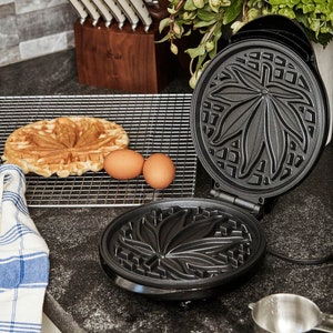 May include: A black waffle maker with a leaf design. The waffle maker is open and a waffle is on a wire rack next to it. There are two eggs on the rack as well.