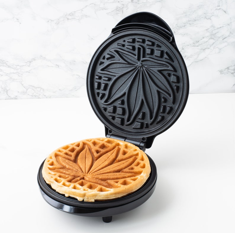 May include: A black waffle maker with a leaf design on the cooking surface. A waffle with a leaf design is in the waffle maker.