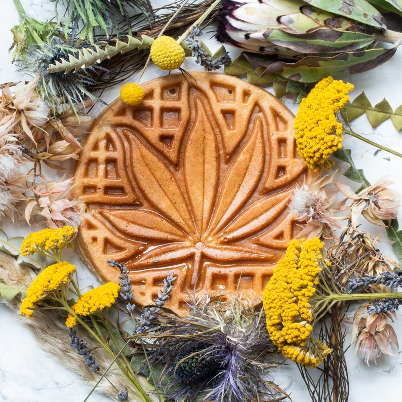 May include: A round, brown waffle with a leaf design. The waffle is surrounded by dried flowers and plants.