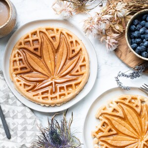 May include: Two golden brown waffles with a maple leaf design on top. The waffles are on white plates and a white surface.