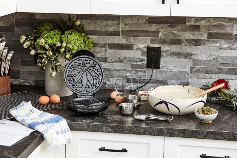 May include: A black waffle iron with a marijuana leaf design on the top plate. The waffle iron is open and there are two eggs, a measuring cup, and a bowl of batter on the counter next to it.