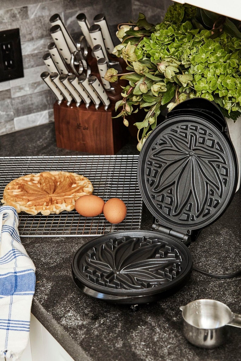 May include: A black waffle maker with a leaf design. The waffle maker is open and a waffle is on a wire rack next to it. There are two eggs on the rack as well.