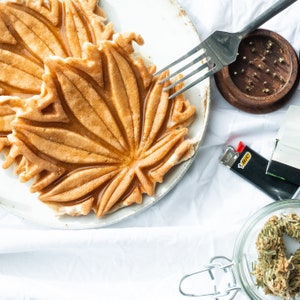 May include: A close-up of a maple leaf-shaped waffle on a white plate. The waffle is golden brown and has a glossy finish. There is a fork and a grinder in the background.