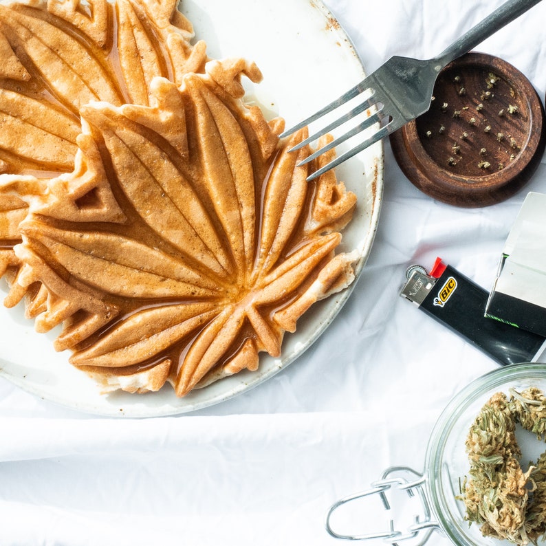 May include: A close-up of a maple leaf-shaped waffle on a white plate. The waffle is golden brown and has a glossy finish. There is a fork and a grinder in the background.