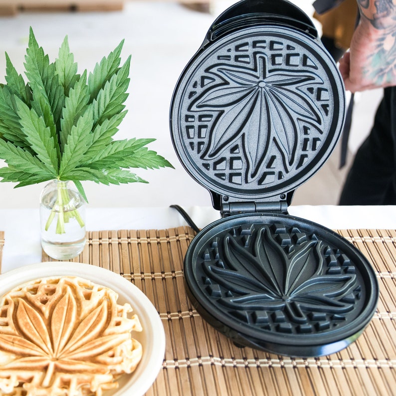 May include: A black waffle maker with a leaf design on the cooking plates. The waffle maker is open, revealing the waffle design. A waffle is being made in the waffle maker.