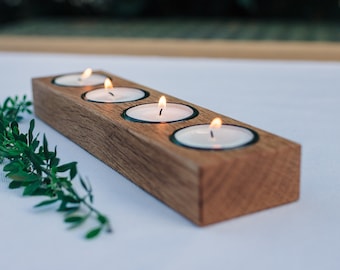 May include: A wooden candle holder with four slots for tea lights. The holder is made of natural wood and has a rustic look. The tea lights are lit and casting a warm glow.