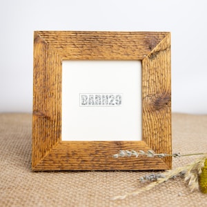 Könnte beinhalten: Ein rustikaler Holzbilderrahmen mit einer natürlichen Holzoptik. Der Rahmen ist quadratisch und hat ein schlichtes Design. Der Rahmen steht auf einer Jutestoff-Oberfläche mit getrockneten Blumen im Vordergrund. Der Rahmen hat einen weißen Passepartout und der Text "BARN29" ist in der Mitte des Passepartouts gedruckt.