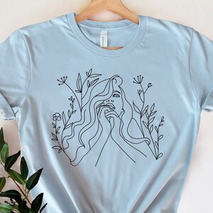 May include: Pale blue t-shirt with a black line drawing of a woman's face and hair, with floral accents. The shirt is hanging on a wooden hanger. The design is a minimalist style.