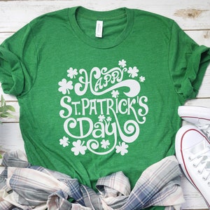 Op de afbeelding: Een groen t-shirt met de witte tekst "Happy St. Patrick's Day" en klaveraccenten. Het shirt wordt gecombineerd met een geruit overhemd en witte canvas sneakers op een wit houten oppervlak.