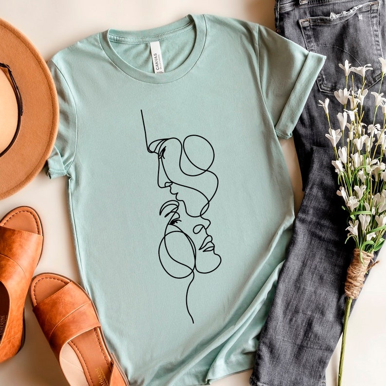 May include: A light blue t-shirt featuring a black line drawing of two faces intertwined.