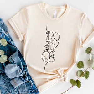 May include: A beige t-shirt featuring a black line drawing of two faces intertwined. The faces are drawn in a minimalist style.