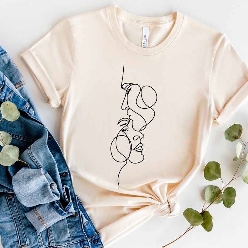 May include: A beige t-shirt featuring a black line drawing of two faces intertwined. The faces are drawn in a minimalist style.